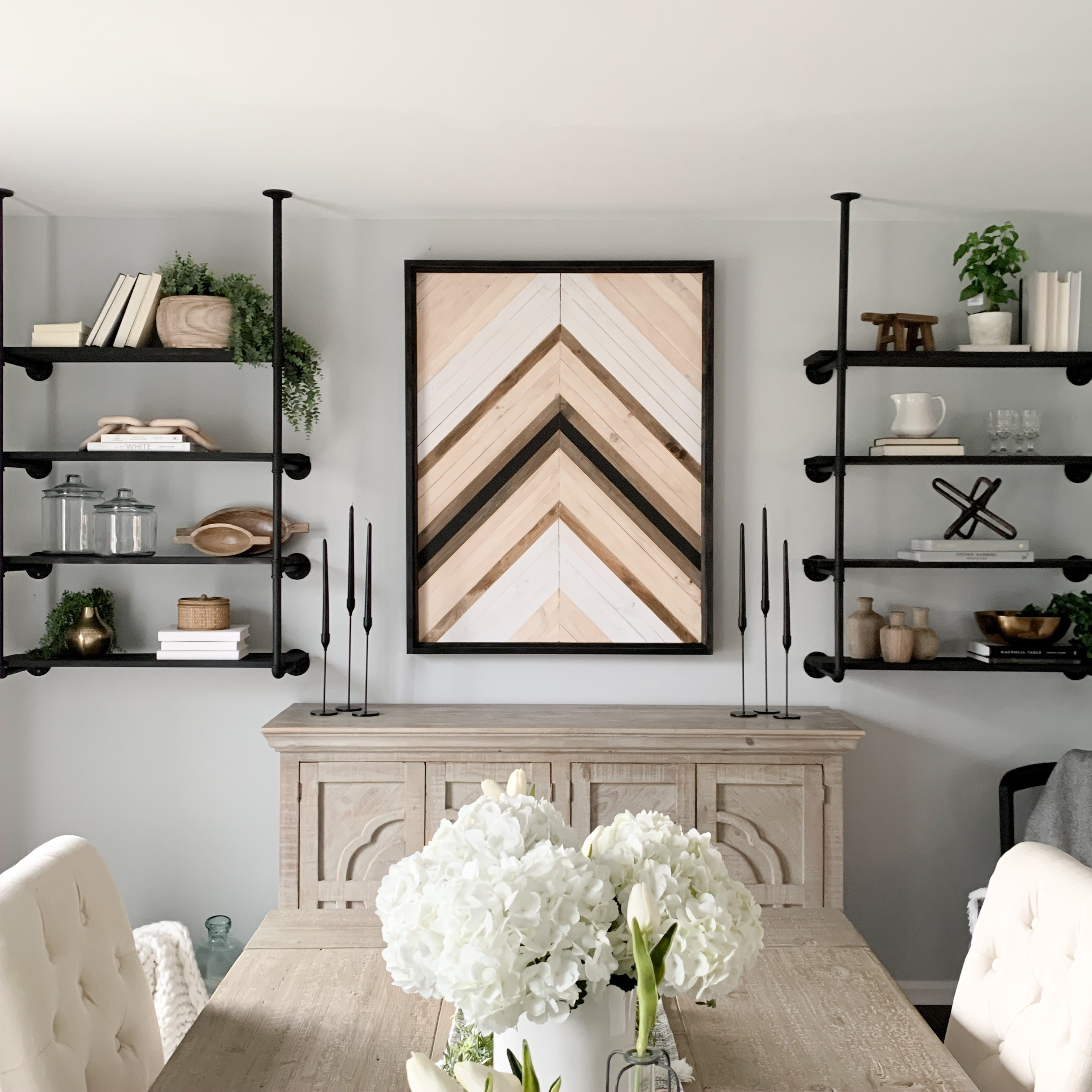 Two sets of affordable DIY industrial pipe shelves installed on wall of dining room with wooden chevron wall art between them.