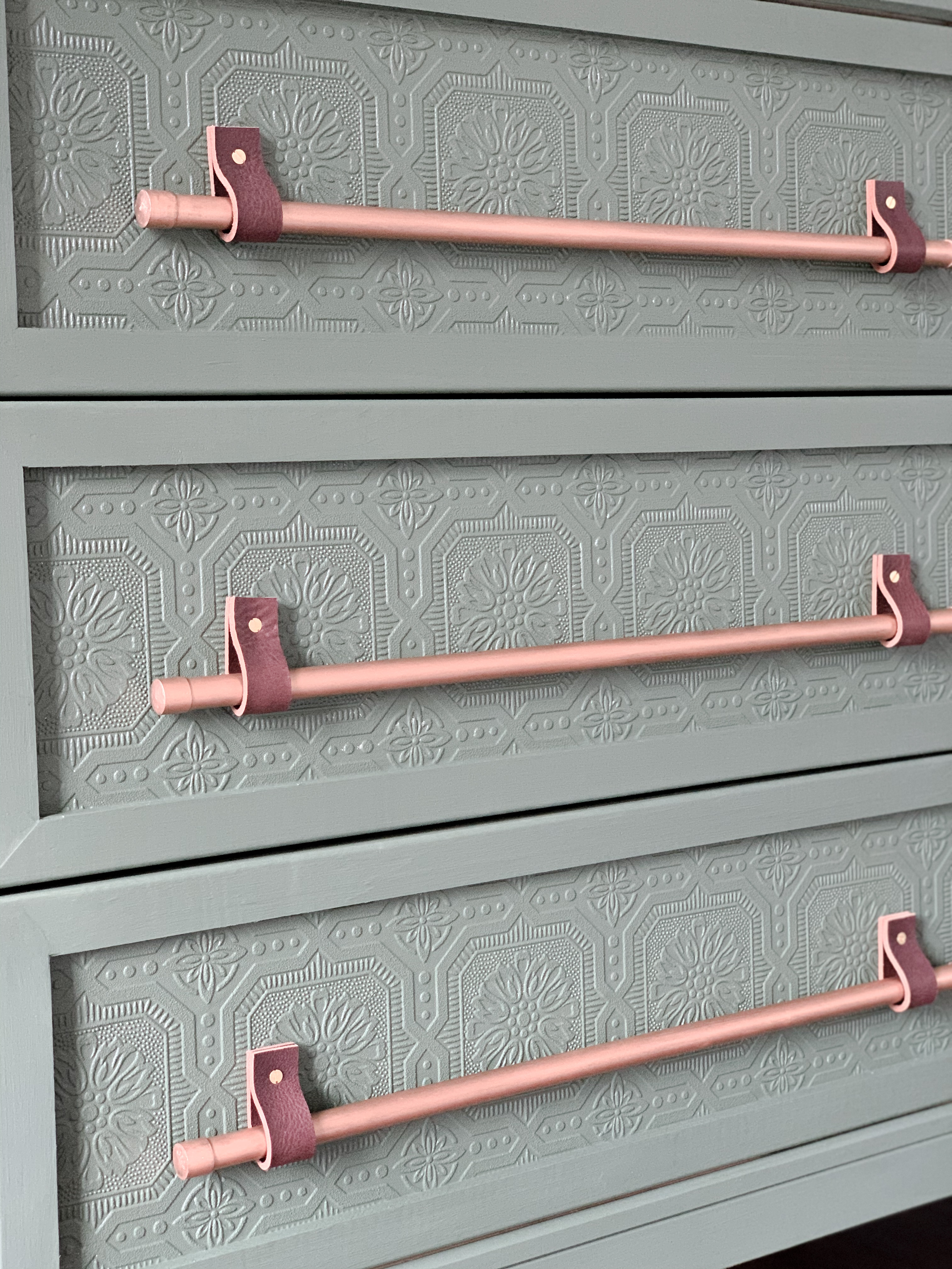 Closeup picture of drawers with green painted wallpaper and copper rods attached with leather straps.
