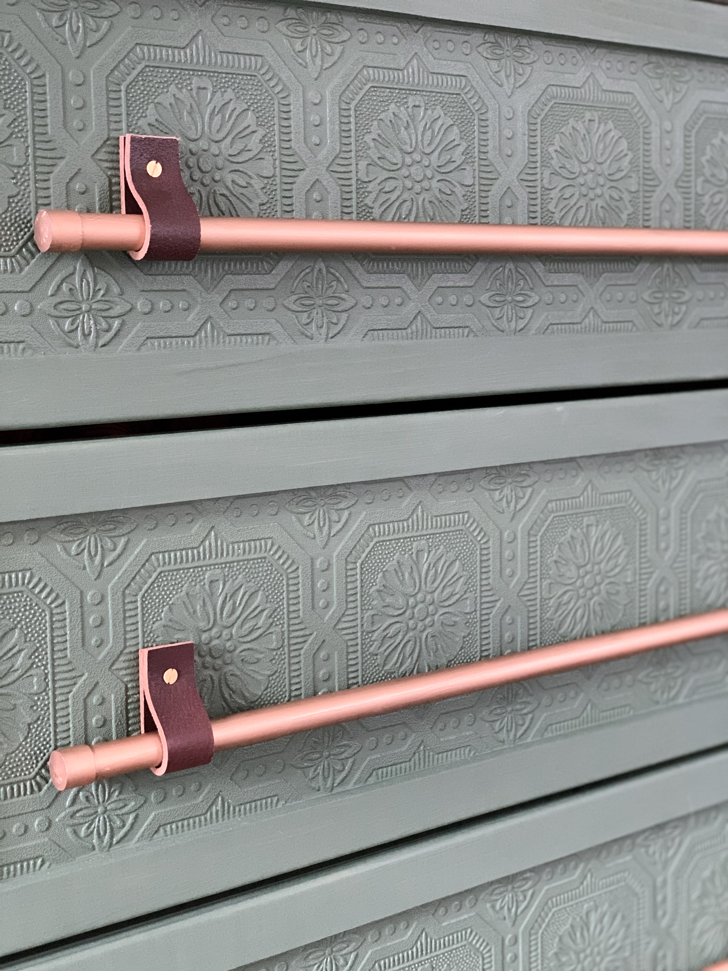 Closeup picture of drawers with green painted wallpaper and copper rods attached with leather straps.
