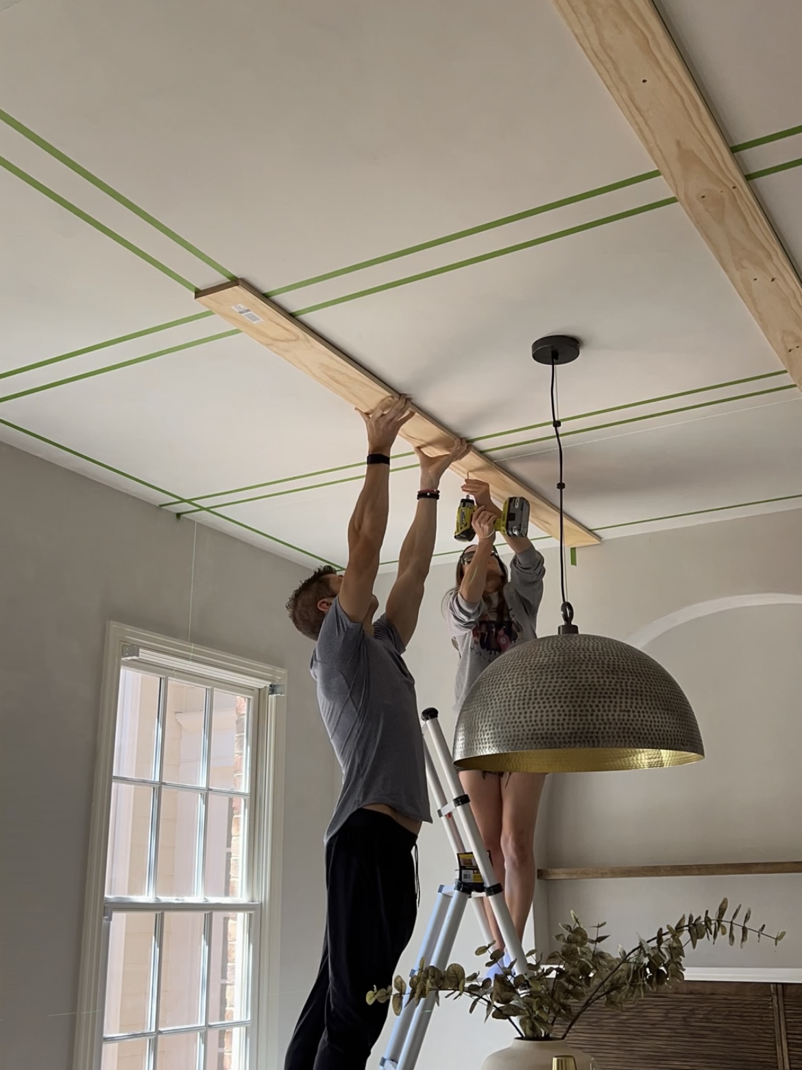 Man holds a board at the ceiling while woman uses a drill to attach the board to the ceiling.