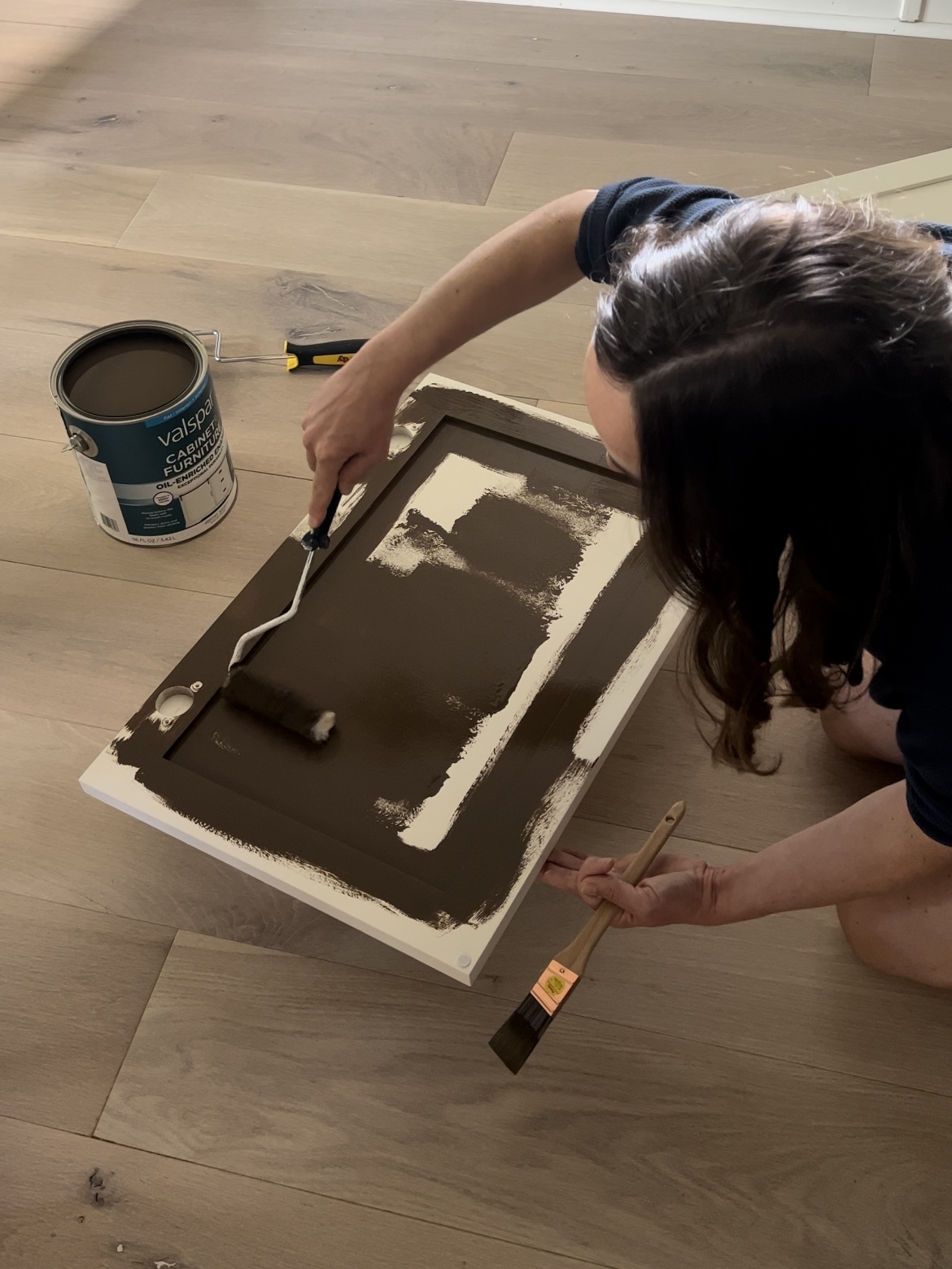 Woman painting a cabinet door brown with a roller and paintbrush.