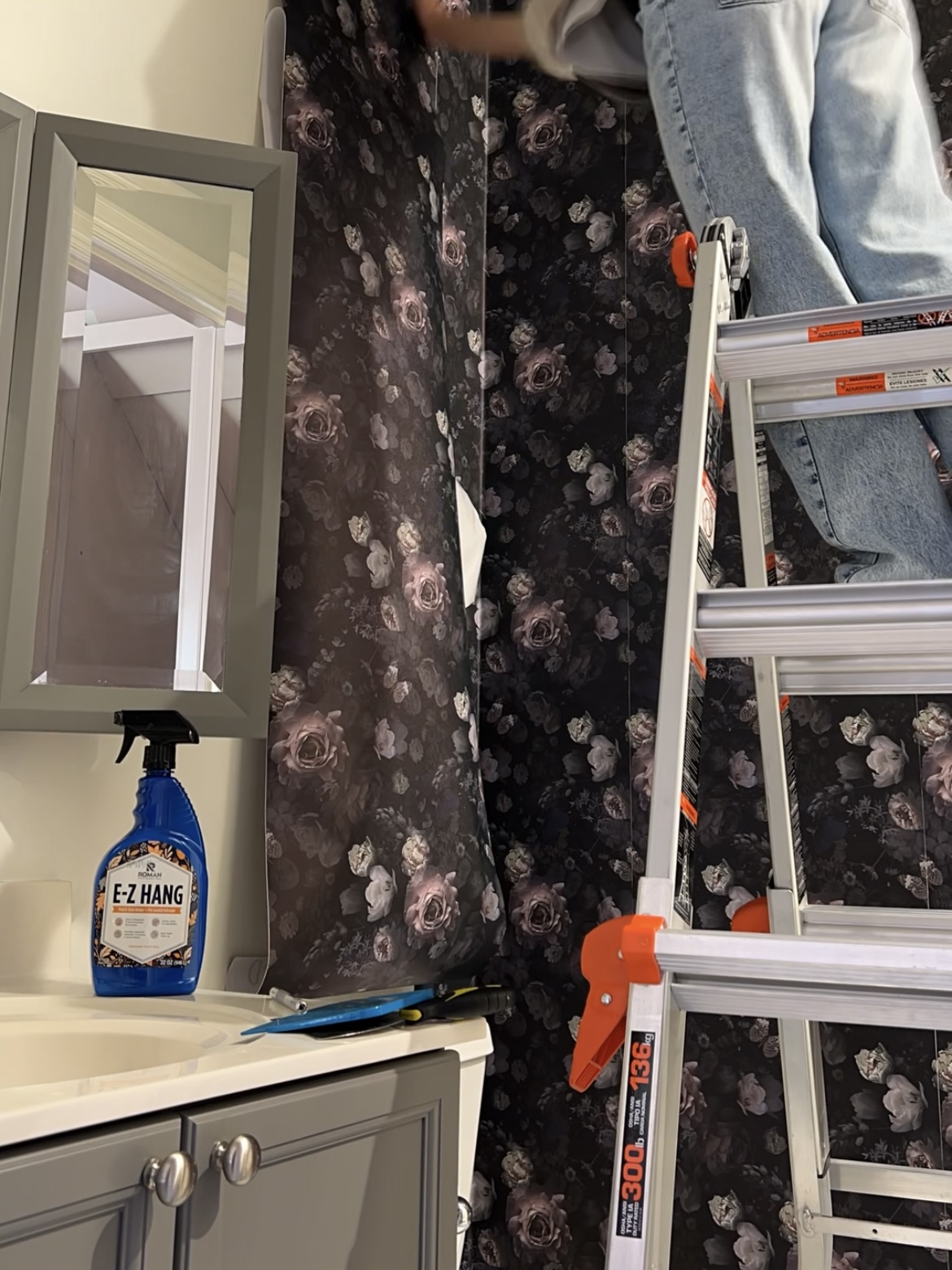 Person on ladder installing floral wallpaper. A bottle of E-Z Hang sits on the counter.