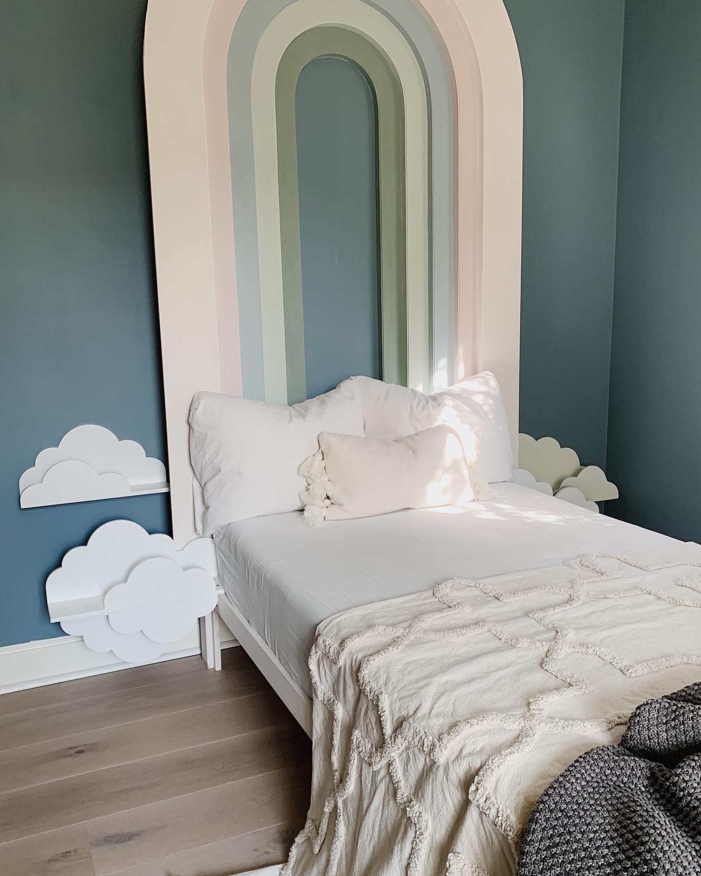 Pastel DIY rainbow headboard above white twin sized bed, with white puffy cloud shelves at bed side level. The bed is made up with white sheets, and has a textural white throw blanket on the end.